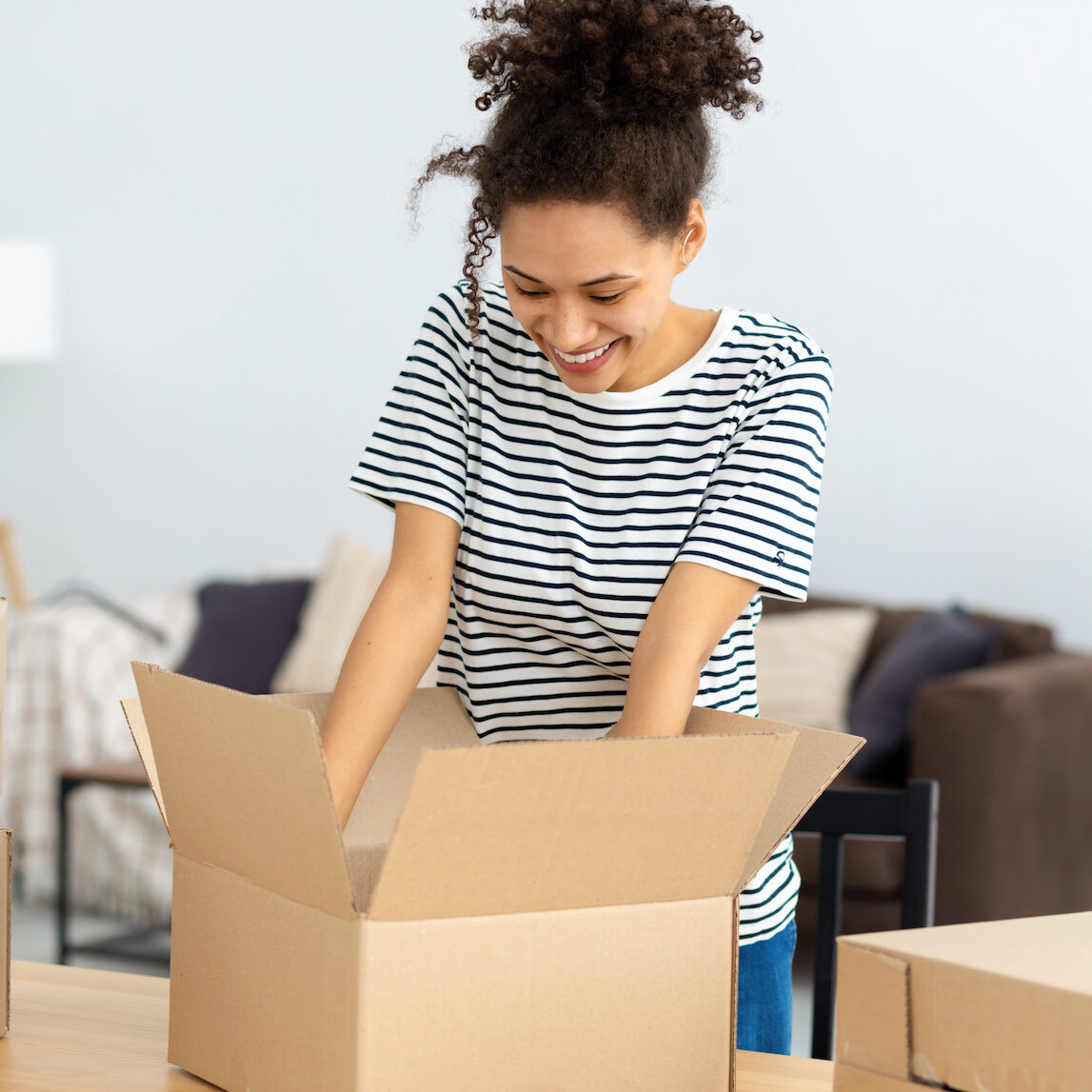 Young smiling African American woman unpacking boxes standing at home. Unpacking parcel, delivery, moving to new house concept. Female Student moves to new home
